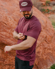 Man wearing a maroon shirt and cap with 'TRWR' logo against a rocky background