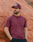 Man wearing a maroon shirt and cap standing against a rocky background