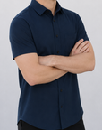 A man wearing a dark navy blue short-sleeve button-down shirt and black pants, standing with arms crossed against a plain light gray wall.