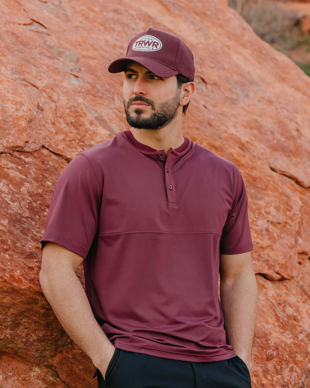Man wearing a maroon shirt and cap standing against a rocky background
