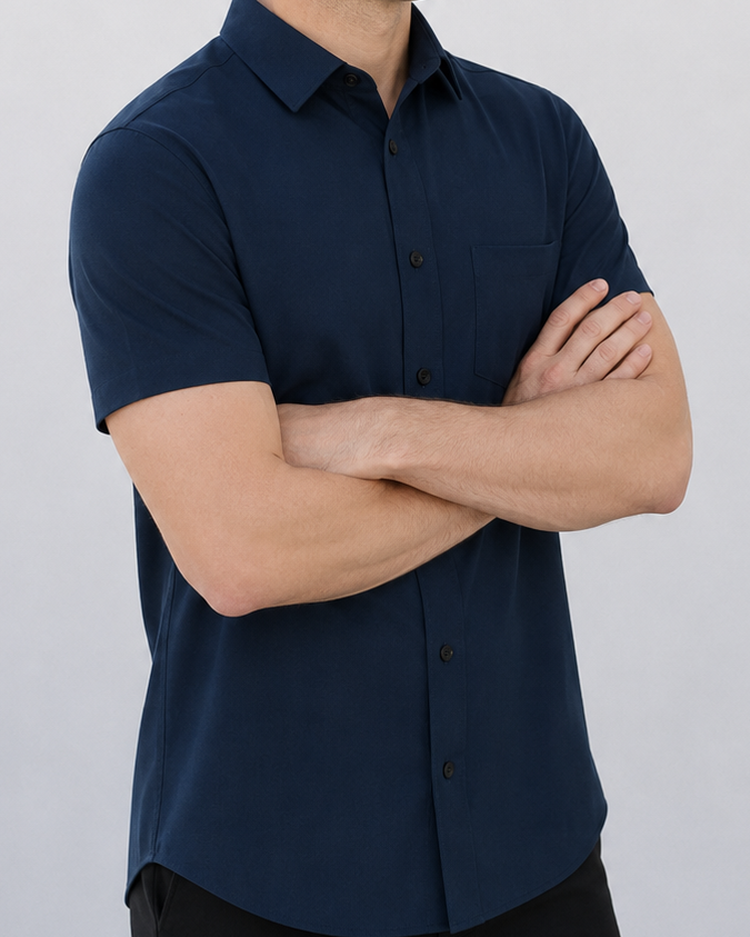 A man wearing a dark navy blue short-sleeve button-down shirt and black pants, standing with arms crossed against a plain light gray wall.